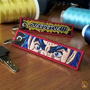 May include: Two embroidered keychains. One keychain is red with a black background and features a close-up of a blue-haired anime character's eyes. The other keychain is yellow with a black background and says "OVERDRIVE!!!".
