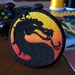 Puede incluir: Un parche bordado negro con una silueta de dragón sobre un fondo de puesta de sol amarillo, naranja y rojo.