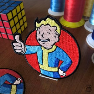 Puede incluir: Un parche bordado rojo, blanco y azul con un personaje de dibujos animados con un mono amarillo, una camisa azul y un pulgar hacia arriba. El personaje sonríe y tiene el pelo rubio. El parche es circular y tiene un fondo rojo.