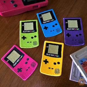 Può includere: Cinque toppe ricamate con il logo di Game Boy Color in diversi colori: verde, blu, viola, giallo e rosa. Ogni toppa ha il testo "GAME BOY COLOR" e un'immagine stilizzata della console Game Boy Color.