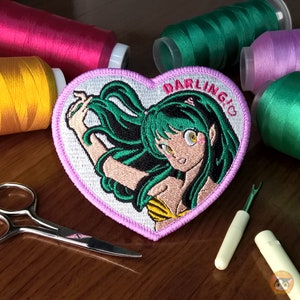 Puede incluir: Un parche bordado en forma de corazón rosa con una chica de dibujos animados de anime con cabello verde y un traje de baño amarillo y negro. El texto "DARLING" está bordado en rosa en el parche.