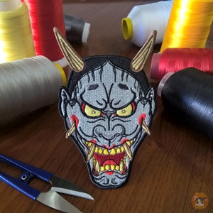 May include: Embroidered patch of a Japanese demon mask with red, yellow, and white details. The mask has a fierce expression with sharp teeth and horns.