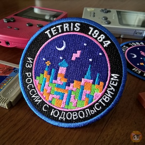 Puede incluir: Un parche redondo bordado en azul y negro con el texto "TETRIS 1984 ИЗ РОССИИ С ЛЮБОВЬЮ" en un círculo blanco sobre un fondo azul oscuro. El parche presenta una ciudad pixelada con una luna creciente y estrellas.