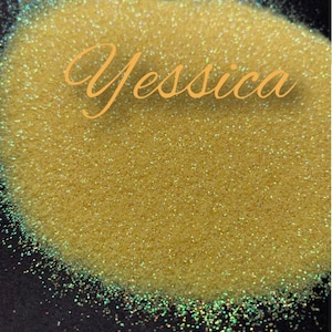 May include: A close-up of a pile of fine, iridescent glitter with a light yellow base colour. The glitter has a subtle shimmer and reflects light in a variety of colours. The word "Yessica" is written in cursive on the glitter.