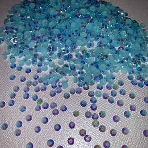 May include: A large pile of light blue iridescent rhinestones, with a few scattered on a white surface.