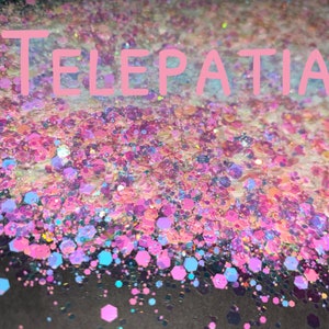 May include: A close-up of iridescent glitter in shades of pink, blue, and white. The glitter is scattered on a black surface and the word "TELEPATIA" is written in pink letters across the top of the image.