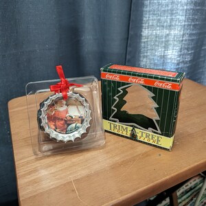 May include: A silver Coca-Cola Christmas ornament featuring a vintage image of Santa Claus. The ornament is packaged in a green box with the Coca-Cola logo and the words "Trim-A-Tree Collection".