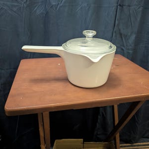 May include: A white saucepan with a long handle and a clear glass lid. The saucepan is sitting on a brown wooden folding table. The background is a dark blue curtain.