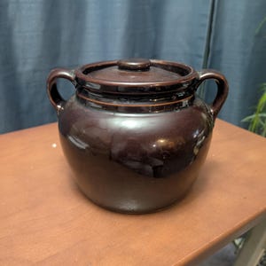 May include: A brown ceramic crock with a lid and two side handles. The crock is sitting on a wooden table. The crock has a glossy finish and a dark brown color.