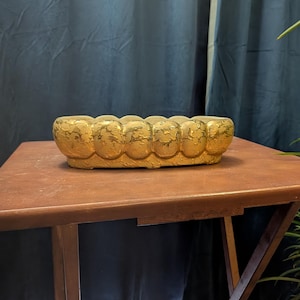 May include: An oval-shaped ceramic planter with a metallic gold finish and a textured, scalloped exterior. The surface features a distressed, crackled pattern throughout the gold glaze, creating a reflective appearance across the rounded edges.