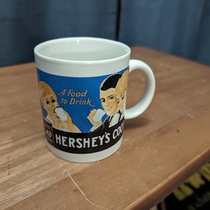 May include: White ceramic mug with a blue and yellow illustration of three people drinking Hershey's Cocoa. The text "A Food to Drink" is printed on the mug.