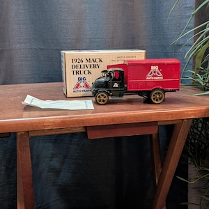 May include: A vintage 1926 Mack Delivery Truck toy with a red cargo bed and black cab, featuring "BIG AUTO PARTS" branding. The toy truck is accompanied by its original box, which reads "LIMITED EDITION COLLECTOR". The toy is on a wooden table.