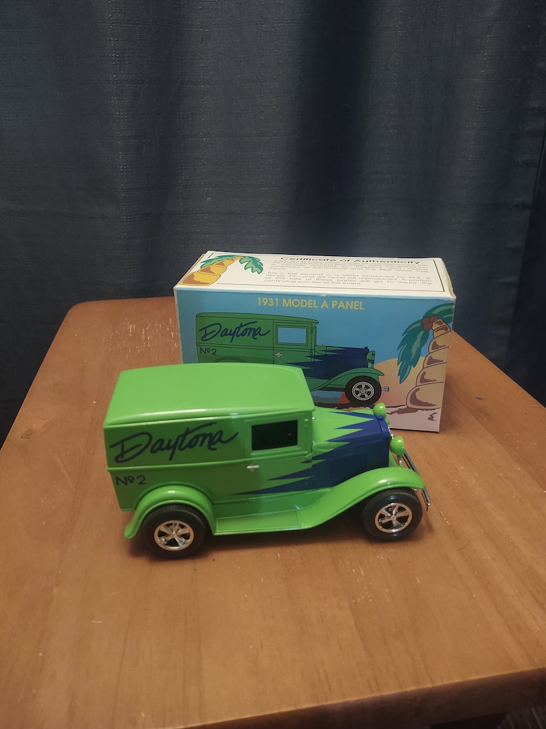 Puede incluir: Un juguete de cami&oacute;n de reparto verde 1931 Model A Panel con una franja azul y "Daytona No. 2" escrito en el lateral. El cami&oacute;n est&aacute; sobre una superficie de madera con una caja detr&aacute;s.