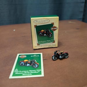 May include: A miniature Harley-Davidson motorcycle ornament, a collector's item, is displayed with its packaging and a small card. The ornament and card feature the text "1933 Flathead Model VLD Miniature Harley-Davidson Motorcycles."
