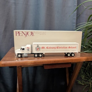 May include: A white and red toy semi-truck model with the text "Mt. Calvary Christian School" on the trailer. The truck is on a wooden table with a box labeled "PENJOY" behind it. The model truck has black wheels.