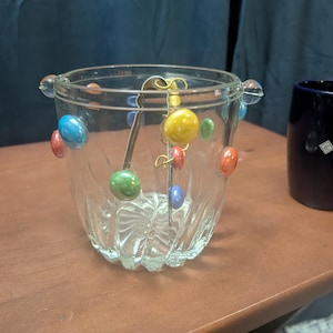 May include: A clear glass ice bucket with colorful, round embellishments. The bucket has a decorative, ridged design and a metal ice spoon. The bucket is decorated with red, blue, green, and yellow accents.