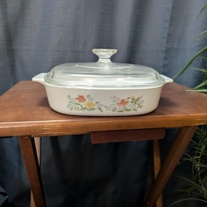 May include: A vintage, rectangular casserole dish with a clear glass lid. The white dish features a floral pattern in shades of orange, yellow, and green. The dish is on a wooden table.