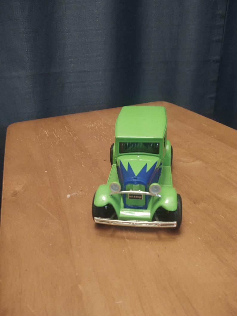 Puede incluir: Un coche de juguete verde con detalles azules y blancos. El coche tiene una parrilla y faros negros. El coche est&aacute; sentado sobre una superficie de madera.