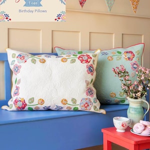 May include: Two floral patterned pillows on a blue bench with a red side table and a floral patterned rug. The pillows are white and light blue with a floral design. The text "Jubilee Tilda Birthday Pillows" is on a sign above the bench.