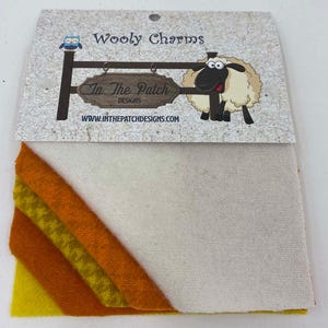 May include: A set of four wool felt squares in shades of yellow, orange, and white. The felt squares are arranged in a diagonal pattern. The packaging includes a label with the text "Wooly Charms" and "In The Patch Designs".