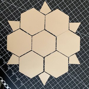 May include: Arrangement of tan-colored geometric shapes, including hexagons and triangles, on a black grid surface. The shapes are arranged in a floral or star-like pattern, with the hexagons forming the central structure.