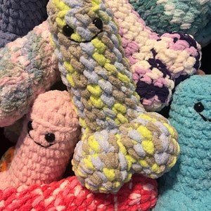 May include: A pile of colorful, plush, phallic-shaped toys. The toys are made of a soft, textured yarn in various colors, including pink, blue, yellow, and purple. Each toy has a smiling face with black button eyes and a stitched mouth.