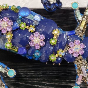 May include: Close-up of a blue, green, and purple mosaic artwork featuring a flower design. The artwork is made of various materials, including glass, beads, and rhinestones.
