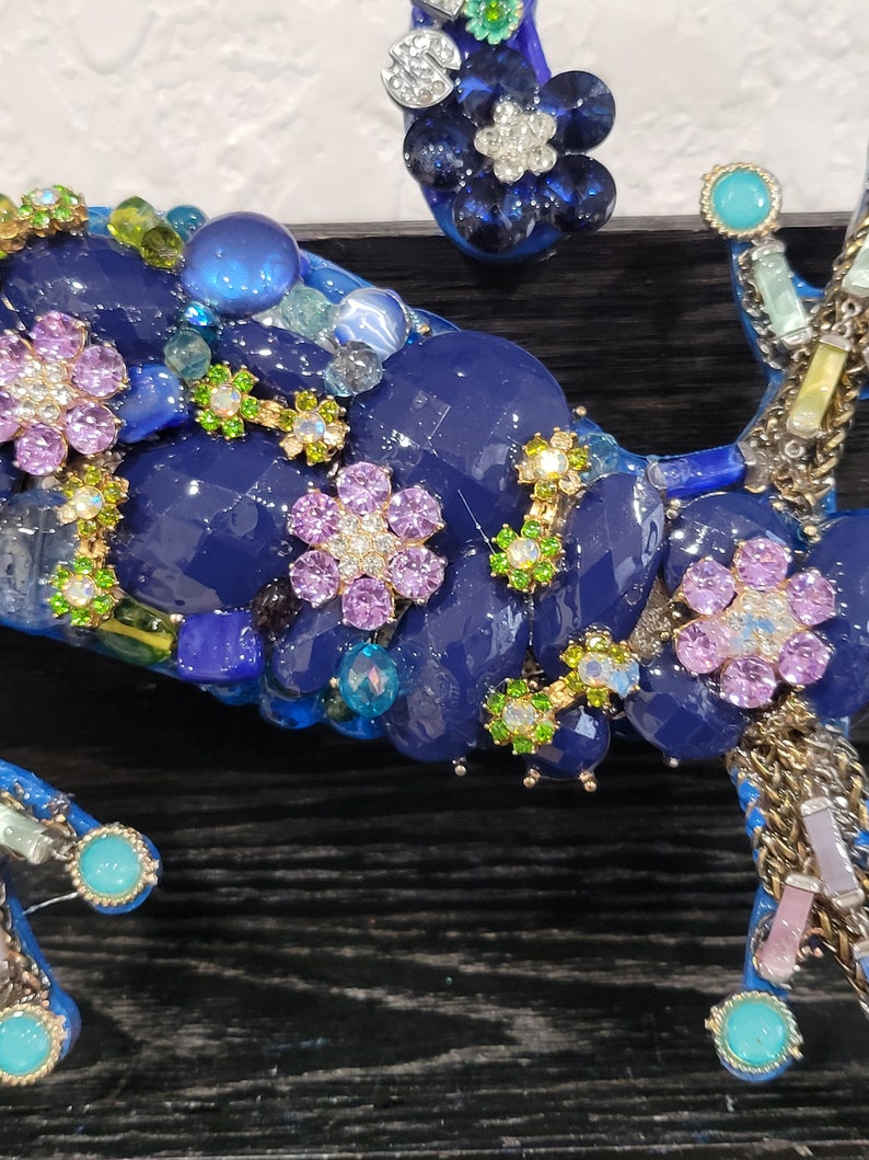 May include: Close-up of a blue, green, and purple mosaic artwork featuring a flower design. The artwork is made of various materials, including glass, beads, and rhinestones.
