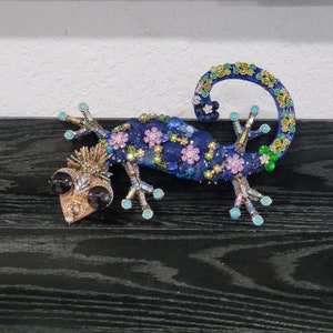 May include: A blue, green, and purple jeweled gecko figurine with black eyes. The gecko is curled up with its tail in the air.