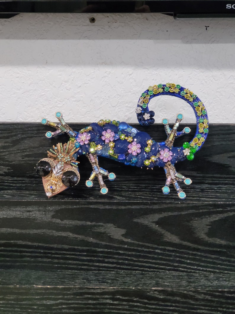 May include: A blue, green, and purple jeweled gecko figurine with black eyes. The gecko is curled up with its tail in the air.