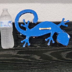 May include: A blue gecko wall decor with a textured finish. The gecko is mounted on a black surface.
