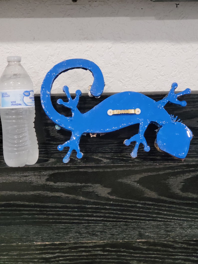 May include: A blue gecko wall decor with a textured finish. The gecko is mounted on a black surface.