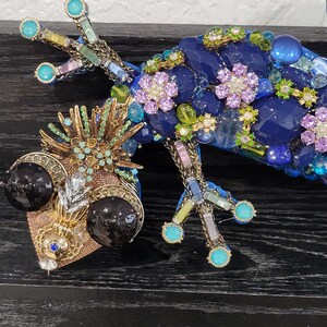 May include: A blue and gold jeweled gecko figurine with black eyes and a detailed design. The gecko is made of glass and metal and is decorated with rhinestones, beads, and other embellishments.