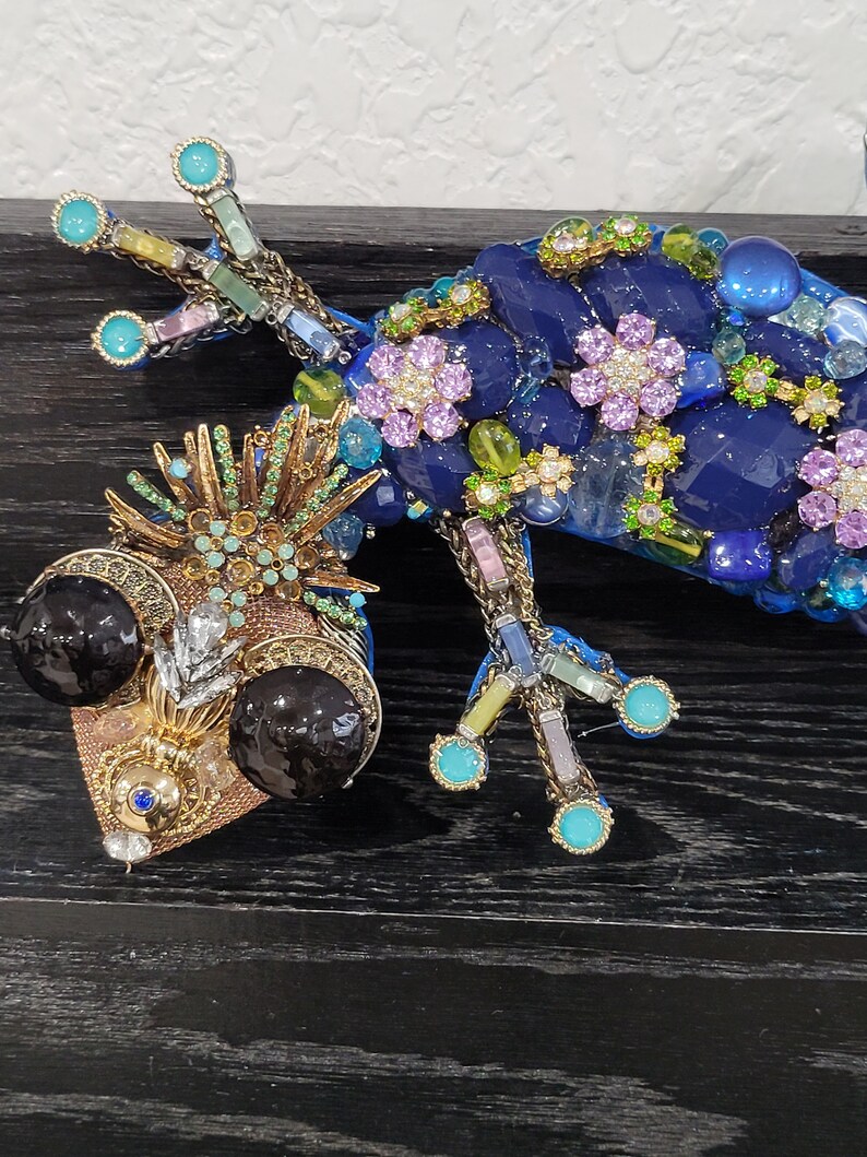 May include: A blue and gold jeweled gecko figurine with black eyes and a detailed design. The gecko is made of glass and metal and is decorated with rhinestones, beads, and other embellishments.