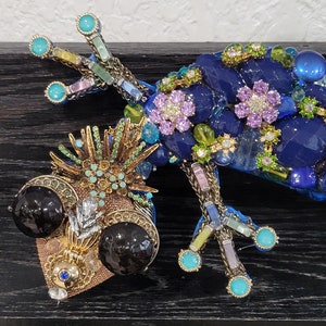 May include: A blue, green, and gold jeweled gecko sculpture with large black eyes. The gecko is made of glass and metal and has a detailed design.