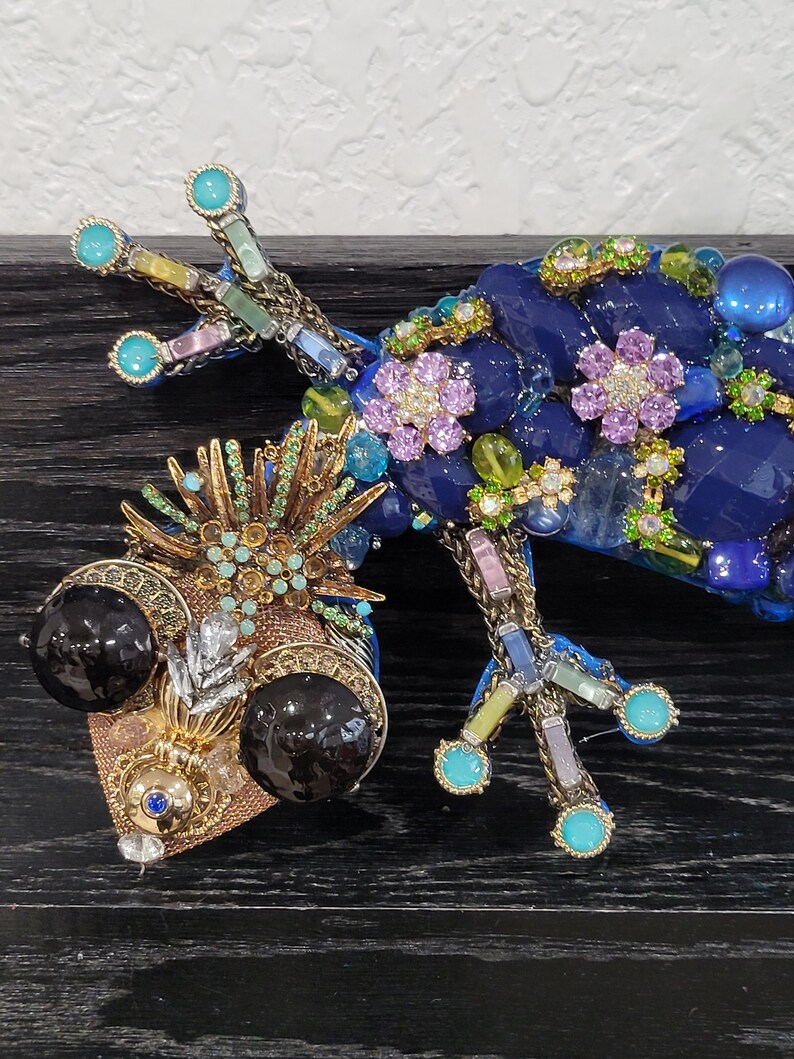 May include: A blue, green, and gold jeweled gecko sculpture with large black eyes. The gecko is made of glass and metal and has a detailed design.