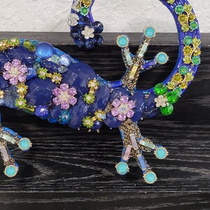 May include: A blue jeweled gecko figurine with green, pink, and white accents. The gecko is curled up with its tail forming a circle.