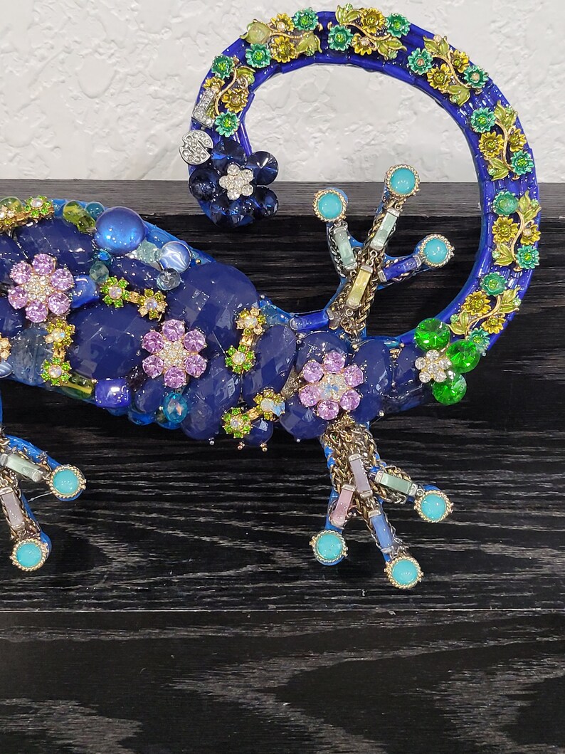 May include: A blue jeweled gecko figurine with green, pink, and white accents. The gecko is curled up with its tail forming a circle.