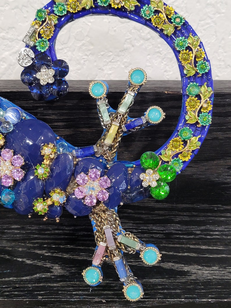 May include: A blue and green mosaic gecko sculpture with colorful flowers and jewels. The gecko is curled up with its tail forming a circle.