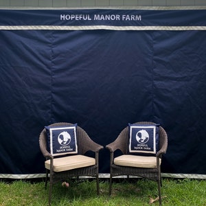 May include: Two wicker chairs with beige cushions and navy blue pillows featuring a white logo and the text "HOPEFUL MANOR FARM." A navy blue backdrop with the same text is behind the chairs. The scene is set on a grassy lawn.