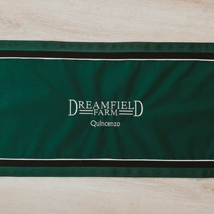 May include: Green fabric banner with white embroidered text that reads "Dreamfield Farm Quincenzo". The banner has black trim and gold metal rings at each corner.