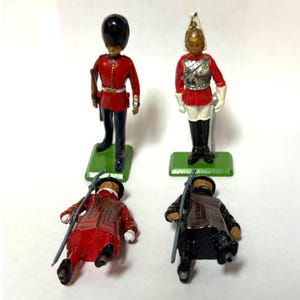 May include: Four metal toy soldier figurines. Two stand upright on green rectangular bases: one in a red tunic with a tall black bearskin hat, and one in a red tunic with a silver breastplate and white helmet. Two lie flat, one red and one black.