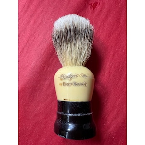 May include: A vintage shaving brush with a cream-colored handle and black base. The brush head features a mix of white and brown bristles. The text "Badger Line by Ever Ready" is printed on the handle.