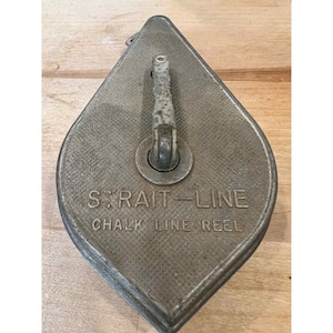 May include: A vintage, teardrop-shaped chalk line reel made of metal. The reel is a dull silver color with the words "STRAIT-LINE CHALK LINE REEL" embossed on the front. It has a handle and a textured surface.