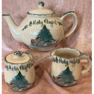 May include: A cream-colored ceramic tea set with a teapot, sugar bowl, and creamer. Each piece is decorated with a Christmas tree, horse, and sheep illustration, and the words "Holy Night". The set has green trim.