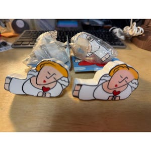 May include: Two identical ceramic or plastic figurines shaped like reclining figures with yellow hair, white clothing, and a small red heart held in their hands. One figure is unwrapped, while the other remains partially inside a clear plastic bag.
