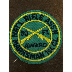 May include: A green and yellow embroidered patch with the text "NAT'L RIFLE ASSN" and "MARKSMAN 1ST". The patch features two crossed rifles and the text "50 FT AWARD".