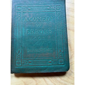 May include: A vintage, teal-colored book titled "A Comedy of Errors" by Shakespeare. The title and author's name are embossed in gold lettering on the cover. The book has decorative borders and a worn appearance, suggesting its age.