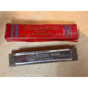 May include: A silver-colored metal harmonica with the text Marine Band M. Hohner engraved on the front, accompanied by a rectangular red cardboard box featuring blue vertical stripes and the text Marine Band No. 365 Made by M. Hohner Germany.