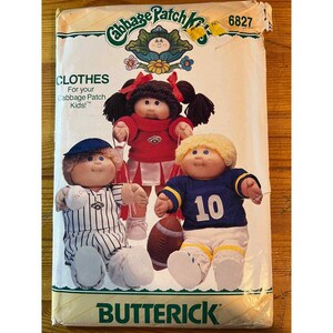 May include: A vintage Butterick pattern for Cabbage Patch Kids clothes. The pattern features three dolls dressed in different outfits: a cheerleader, a baseball player, and a football player. The pattern's packaging is cream-colored with the Cabbage Patch Kids logo.